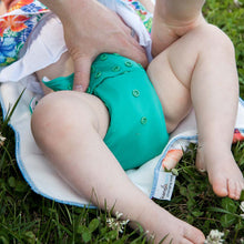 Load image into Gallery viewer, putting a Peacock Rumparooz on a baby who is laying on a changing pad on the grass