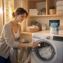 Load image into Gallery viewer, Woman loading a washing machine with a container of Mighty Bubbles laundry treatment in the foreground.